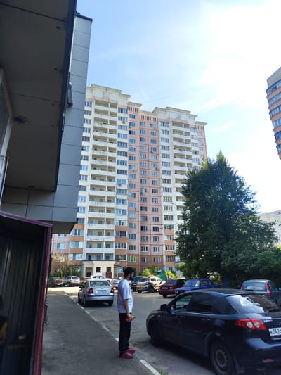 A man in a white t-shirt and black pants stands on a paved surface, holding a red object in his right hand. He is facing to the left. Behind him, several parked cars are visible. In the background, a large, tall apartment building with numerous balconies and windows dominates the scene. A dense green tree is located to the right of the building. The sky is clear and blue, with some faint cloud streaks. The setting appears to be an urban environment, likely in the city of Zhukovsky, Russia, given the context provided.