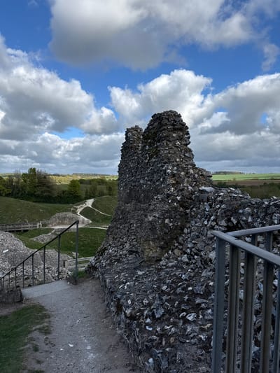 The image presents a panoramic view from a vantage point atop a historical ruin, likely a castle or fortified wall, under a dynamic sky. The scene is dominated by a substantial, weathered stone wall constructed from countless irregular grey and white flint or chalk fragments, giving it a rugged, textured appearance. This ancient masonry forms the right and lower portions of the frame.
Above and to the left of the wall, a bright blue sky is interspersed with large, white, and grey cumulus clouds, indicating partially cloudy weather during daytime. Beyond the immediate structure, a vast, undulating green landscape unfolds, featuring cultivated fields, scattered trees, and distant horizons. Winding paths or tracks are visible on the grassy slopes below the ruins, suggesting areas for walking or exploration within a historic site.
Several metal railings are incorporated into the scene, appearing as safety barriers or part of a visitor access structure, offering a sense of scale and indicating public accessibility to the ruins. The general activity implied is sightseeing or exploring a heritage location.
The time of day appears to be bright daylight, possibly morning or afternoon. The characteristic flint construction and rolling agricultural landscape are consistent with the provided location context of King's Lynn and West Norfolk, United Kingdom, which is known for its historical sites and chalk/flint geology. No discernible text is visible on any signs or objects within the image.