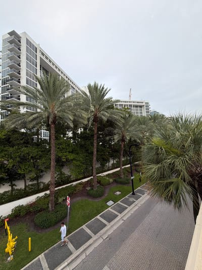 A high-angle view captures a paved walkway alongside a grassy area with several palm trees in Bal Harbour Village, Florida, USA. A man in a white long-sleeved shirt and patterned shorts walks along the walkway. A red "Do Not Enter" sign is visible on a pole in the grassy area, along with a yellow sign and yellow pole further to the left. Behind the greenery and palm trees, a modern white building with multiple balconies and large windows stands tall, its structure stretching horizontally across the frame. The sky above is overcast.