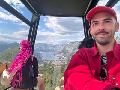 A medium eye-level shot captures three individuals inside a cable car, ascending over the city of Mirac, Montenegro. The main subject is a fair-skinned man with a beard, a red shirt, and a matching red baseball cap. His sunglasses hang from his shirt's collar. To his left is a woman with vibrant pink braids, wearing a black dress, with a beige bag resting beside her. Another person with dark hair is seated partially obscured behind them. Through the cable car's window, there's a vista of the city, a bay filled with boats, and a mountainous backdrop. The sky is partly cloudy. The cabin features a black metal frame and a security camera on the ceiling.