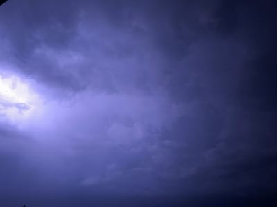 The image captures a dramatic sky over Świdnica, Poland, dominated by dark, heavy storm clouds. A faint, bright lightning strike is visible on the left side of the frame, illuminating the turbulent cloud formations with a purplish-blue hue. The lower portion of the image shows a silhouetted figure against the dark horizon, suggesting the presence of a person observing the storm. The overall scene evokes a sense of impending weather and the raw power of nature.