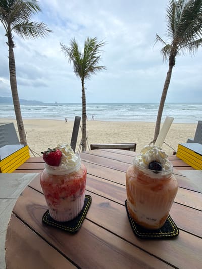 The image depicts a static, eye-level view from a beachside establishment in An Hải Ward, Vietnam, on an overcast day. In the immediate foreground, a light brown, slatted wooden table holds two drinks. The drink on the left is a layered pink and red smoothie contained within a clear, ribbed glass jar, topped with white whipped cream and a single whole red strawberry. A dark grey straw is inserted into this drink. The drink on the right is a layered orange and peach smoothie, also in a clear, ribbed glass jar, topped with white whipped cream and a single dark berry, likely a blueberry. A light-colored straw is inserted into this second drink. Both jars rest on black square coasters with prominent yellow stitching.

Beyond the table, a wide expanse of sandy beach stretches towards the ocean. The ocean features moderate waves breaking near the shore. Three tall, slender palm trees with green fronds are spaced across the midground, their trunks providing vertical elements. At least two individuals are observed walking on the sandy beach, one positioned to the left and another to the right of the central view, both appearing to walk parallel to the shoreline or towards the water. In the far distance, a large vessel, consistent with a ship, is visible on the ocean horizon, positioned slightly to the left of the image center. The sky above is largely overcast with a mix of white and grey clouds. Faint outlines of distant mountains or hills are discernible on the far left horizon. Partial views of grey and yellow-accented seating or structural elements are visible on the left and right sides of the foreground.