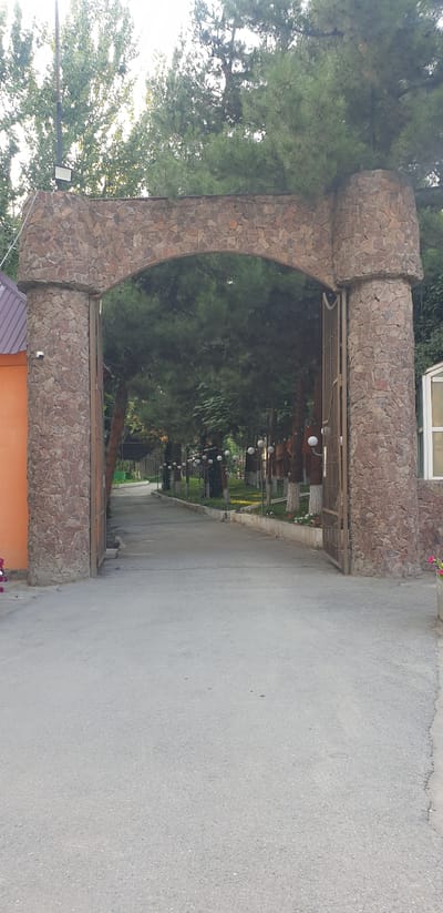 The image shows a large arched gateway made of rough-hewn stones. The gateway is open, revealing a paved pathway leading into a tree-lined area with decorative lampposts. To the left of the gateway is an orange building with a pitched roof and a security camera mounted on the stone pillar. To the right, a portion of a building with a window is visible. The pathway is bordered by a low concrete planter with some small flowers. The scene appears to be an entrance to a park, garden, or possibly a residential complex in Avval', Uzbekistan, given the style of architecture and vegetation. The lighting suggests it is daytime, possibly late afternoon or early morning, with the sun casting a soft light. There are no people visible in the image.