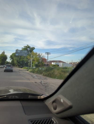 The image depicts a view from inside a car traveling on a paved road in Beş-Küŋgöy, Kyrgyzstan. The sky is visible through the windshield with power lines running across it. To the right of the road is overgrown foliage and dilapidated buildings. There are vehicles in front of the car. The interior of the car, including the interior trim and air vent, is in the foreground.