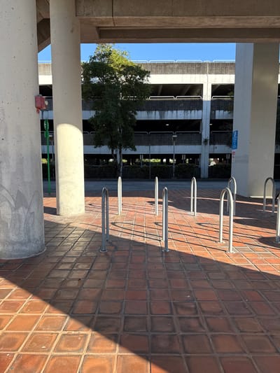 A sideways view shows a parking garage framed by concrete structures. A tree stands between the garage and the columns. To the right, a row of metal bike racks casts long shadows on the brick-paved ground. The sunlight creates a sharp contrast of light and shadow, emphasizing the texture of the brick. The composition is architectural, with a focus on lines and perspective.