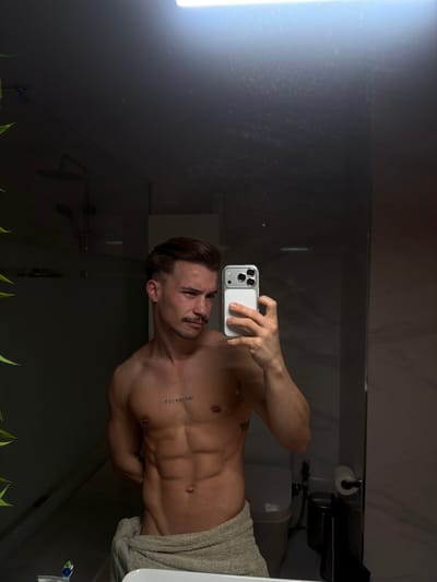 The image displays a male subject taking a mirror selfie in a modern bathroom. The subject, appearing to be in his 20s or 30s, has light brown hair, a mustache, and is shirtless, revealing a muscular torso with visible abdominal muscles, bilateral nipple piercings, and tattoos on his upper left chest (reading "IMPERFECT" vertically) and left rib cage. He is wrapped in a light-colored towel around his waist, holds a light-colored smartphone with a multi-lens camera in his right hand to capture his reflection, and directs his focused gaze towards his right.
The bathroom environment, reflected in the mirror, features a shower area with a rain showerhead and handheld fixture behind a glass partition on the left. To the right, a white toilet with a closed lid is visible, accompanied by a dark waste bin and a wall-mounted toilet paper holder. A toothbrush and toothpaste tube are partially visible on a sink counter in the lower-left foreground, adjacent to green foliage from an unseen plant. An overhead light fixture brightly illuminates the scene from above, causing a prominent reflection on the mirror's surface. The walls appear to be light-colored tile or paneling. No visual information within the image indicates a specific location in Viseu, Portugal.