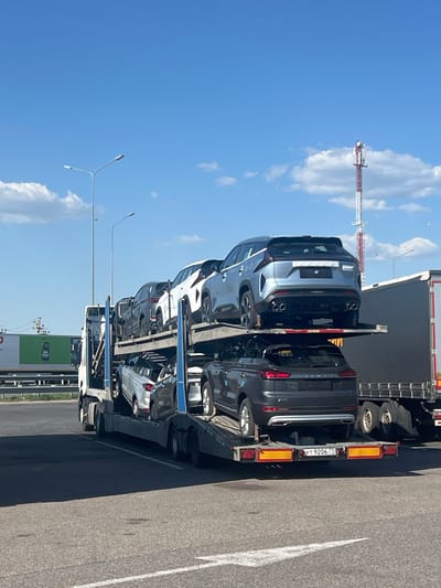 The image shows a car carrier truck loaded with new cars, likely in transit, parked on a paved surface alongside a road. The sky is blue with scattered white clouds. A communication tower is visible in the background. The location is Казань, Russia, identifiable by the license plate "YT 9206 77" on the trailer, which corresponds to Russian vehicle registration codes. Streetlights are also present along the road. There is a large green and white building sign near the truck.