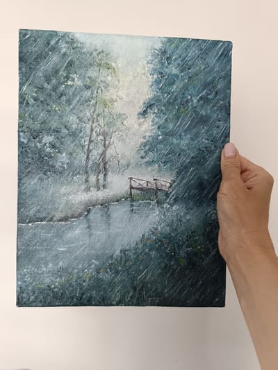 The image shows a painting of a tranquil, rainy forest scene. A small wooden bridge crosses a calm body of water, with trees and foliage lining the banks. The rain is depicted by numerous diagonal white brushstrokes, creating a sense of atmosphere and movement. The overall mood is peaceful and somewhat melancholic, enhanced by the soft, diffused light filtering through the trees and the muted color palette. The painting is held by a person's hand, partially obscuring the right side of the canvas. The background is a plain, light-colored wall. No text is visible in the image. The provided location context is Olsztyn, Poland, but there are no specific visual cues within the painting to confirm this location.