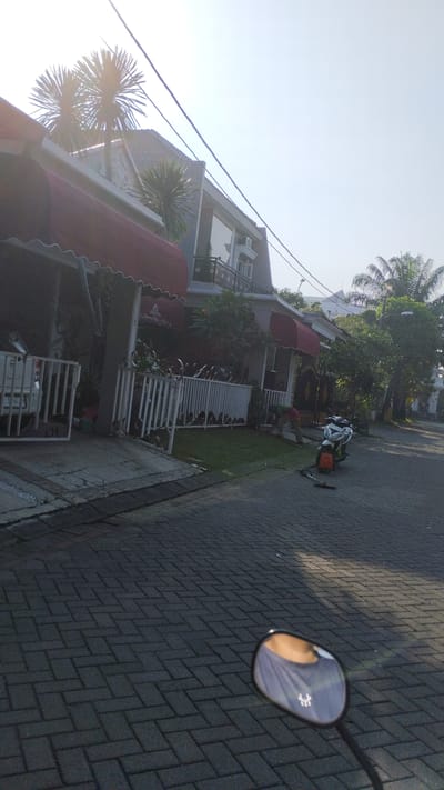In the city of Sidodadi, Indonesia, a residential street is captured on a clear day. The foreground is dominated by a paved walkway, with a motorcycle side mirror reflecting the neck and shoulder of a person wearing a grey t-shirt with a white logo. Beyond the walkway, a white picket fence borders a manicured lawn in front of a modern multi-story house with red awnings over the windows. A person in a green shirt is bent over near the lawn, possibly tending to it. A white pickup truck is parked in front of the house. A scooter is parked on the street. Electrical wires are visible crisscrossing the sky above the houses and trees. Palm trees and other lush vegetation surround the property, suggesting a tropical or subtropical climate. The scene conveys a sense of peaceful, suburban life in Indonesia.