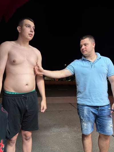 Two young men are standing outdoors at night. The man on the left is shirtless and wearing black shorts with a green waistband. He is looking to his left. The man on the right is wearing a light blue polo shirt and distressed denim shorts. He is reaching out and touching the chest of the man on the left. They are standing on asphalt, and in the background, there is a street with some lights and vegetation. The scene appears to be in Giurgiu, Romania.
