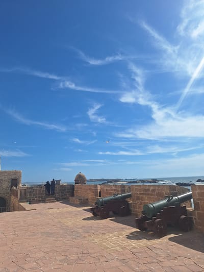 The image shows a section of the ramparts of Essaouira, Morocco, overlooking the Atlantic Ocean. Two antique cannons are positioned on a stone wall, pointing towards the sea. The ground is paved with red cobblestones, and the walls are constructed of weathered brick. Two figures, one wearing a dark hooded garment and the other in darker clothing, stand near a parapet on the left side of the image. In the distance, the ocean stretches to the horizon, dotted with rocky islets. The sky is a clear blue, with wispy white clouds and a bright sun casting shadows on the scene. Seagulls are visible in the sky and near the water. The architecture suggests a historical fortification, likely part of the city's defensive walls.