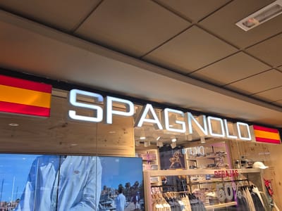 The image shows the exterior of a clothing store in Collado Villalba, Spain. The store's name, "SPAGNOLO," is displayed in large, illuminated white letters across the storefront. A Spanish flag is visible to the right of the sign. Below the sign, a large digital screen displays a scene of people near a body of water, possibly a marina. The store interior, visible through the glass, contains shelves stocked with clothing and shoes, and racks of hanging garments. A sign indicating "REBAJAS" (sales) and "50%" is also visible inside. The overall lighting suggests it is daytime.