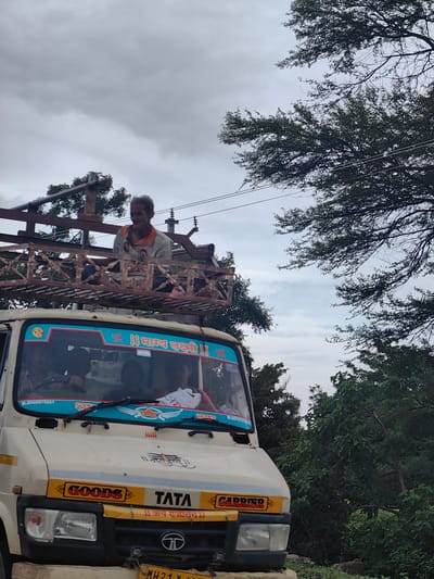 A man is seated on top of a white Tata truck, which is adorned with religious decals. The truck is on a road with trees on either side, under a cloudy sky. The man appears to be a passenger or worker on the truck. The scene is in Walsawangi, India.
