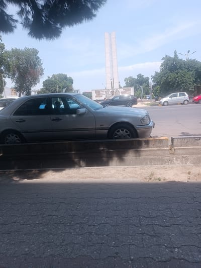 The image depicts a street scene in Khujand, Tajikistan, on a sunny day with a clear sky. In the foreground, a silver sedan is parked partially on a concrete curb. The car's side profile is visible, showing its doors, windows, and wheels. The curb is weathered and has some vegetation growing out of it. Beyond the car, a paved road stretches into the distance.
In the middle ground, several other vehicles are present, including a silver hatchback and a red sedan. A large, modern-looking monument, possibly a memorial or landmark, stands tall in the background, flanked by trees. The monument appears to be made of light-colored stone. The environment suggests an urban setting with signs of everyday city life. The overall impression is a typical street view in a populated area.