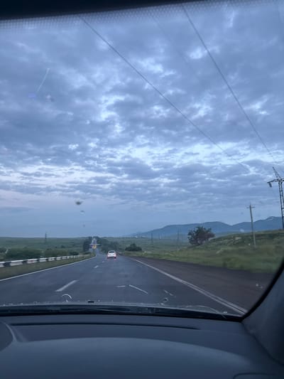 The image depicts a view from inside a vehicle traveling on a highway towards Hrazdan, Armenia. The sky is overcast with dramatic clouds, suggesting either early morning or late evening. The highway is asphalt, with white lane markings visible. In the distance, a white car is driving ahead. The surrounding landscape is characterized by rolling hills covered in green grass and sparse vegetation, with several power lines and transmission towers dotting the scenery. Mountains are visible in the far background, partially obscured by the atmospheric conditions. The windshield of the vehicle shows some smudges and debris, and a Mercedes-Benz emblem is visible on the hood. The overall impression is one of a journey through a rural, possibly mountainous, region under a moody sky.