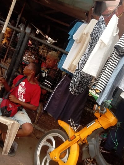 The image captures an outdoor market scene in Umudioka, Nigeria. In the left foreground, a male individual is seated on a white plastic stool. He wears a red short-sleeved t-shirt with a white graphic print, light-colored shorts, and dark footwear. A red shoulder bag with black straps is positioned across his chest. His head is turned towards the upper right portion of the frame, and his right hand is slightly raised.
Behind the seated male, a dark metal grid structure is visible, likely forming part of a stall. Behind this grid, a second individual is partially visible, characterized by short, light-colored hair and a dark top. This person appears to be observing the scene. Further obscured figures and items are present in the deeper background to the left.
On the right side of the frame, a variety of short-sleeved shirts are displayed vertically on hangers or a line. These include items in light blue, cream, white with black animal print, plain white, and black-and-white stripes. Below these, a pair of dark shorts with a white stripe along the side is also displayed.
In the right foreground, the front portion of an orange/yellow moped or scooter is prominent. It features a bright orange front fender, handlebars, and a silver-spoked front wheel. A small chrome rack is mounted above the front fender. The overhead structure includes visible wooden beams and corrugated material, indicative of a market stall's roof. The ground surface is dark.
