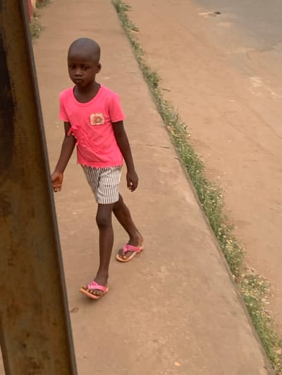 A child wearing a pink t-shirt, striped shorts, and pink sandals stands on a concrete surface near a patch of grass. The child gazes directly at the camera. The shot appears to be taken from an elevated viewpoint, looking down on the child. A dark, blurred object is visible in the lower left corner of the frame. The lighting is soft and muted, suggesting an overcast day.