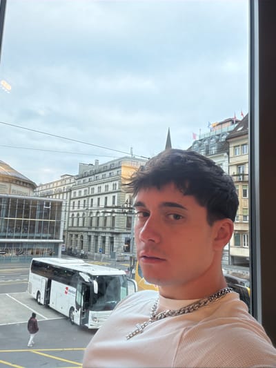 A person, likely male, is shown in a medium close-up view, slightly angled, seemingly from inside a building looking out a window. The person has short dark hair, fair skin, and is wearing a light-colored top and a silver chain necklace with a cross pendant. Beyond the window, the scene is urban, revealing a city street in Lucerne, Switzerland. The architecture is characterized by multi-story buildings with ornate facades. A white bus with the word "maaskant" written on it is parked along the street. A smaller glass building labeled "Panorama" is visible, and a pedestrian can be seen walking on the sidewalk next to the bus. The sky is overcast. Flags are displayed on top of some of the buildings, further indicating the Swiss location.