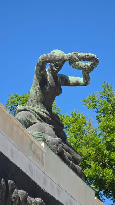 A low-angle, upward-looking shot captures a monumental bronze sculpture of a reclining figure under a clear blue sky. The sculpture, likely weathered bronze with a prominent verdigris patina, depicts a human figure, possibly male, lying on its back. Its left arm is raised, holding a large, detailed wreath aloft. The figure's body is stylized, with visible musculature and flowing drapery.
The sculpture rests on an angled architectural structure, possibly a plinth or part of a larger monument, constructed from light-colored stone or concrete panels. This structure creates a strong diagonal line running from the lower right towards the upper left of the frame. Lush green tree foliage is visible against the bright blue sky, particularly along the top and to the left of the sculpture, suggesting an outdoor setting in a park or public area.
The image conveys a sense of solemnity and remembrance, characteristic of a memorial. The bright, clear blue sky indicates a sunny day. There is no visible text in the image. This sculpture is part of the Monument to the Victims of Fascism in Subotica, Serbia.