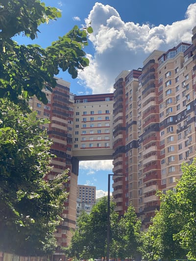 This is a low-angle, vertical shot of modern apartment buildings and lush green trees on a sunny day. The buildings are tall and appear to be new, with a mix of beige, red-brown, and dark gray facades. They feature numerous windows and balconies, suggesting a residential area. Some of the buildings have a skybridge connecting them. The trees, mostly in the foreground and along the right side, are full of vibrant green leaves, casting shadows on the buildings and the ground. The sky is a clear blue, with fluffy white clouds adding depth and texture. The lighting indicates it is daytime, likely late morning or early afternoon, with the sun high in the sky. The scene evokes a sense of urban living integrated with nature, possibly in a city park or a well-landscaped residential complex. The overall mood is bright and cheerful. There is no visible text in the image.