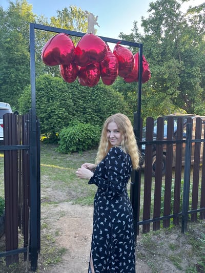 A young woman with long, blonde hair and a black dress with white floral patterns is standing outdoors, smiling towards the camera. She is positioned to the right of a black metal frame from which several red, heart-shaped balloons are hanging on the left side of the frame. The woman is wearing a dark-colored dress with a pattern of light-colored, possibly white or silver, floral or leafy designs. Her hands are clasped in front of her.

The setting appears to be a backyard or outdoor area in Urmanets, Russia, with lush green foliage and trees in the background, suggesting a daytime scene, possibly late afternoon given the lighting. The sky is bright blue and clear in the upper left corner. Behind the woman, there's a dark brown slatted fence, and a dark-colored car is partially visible behind the fence. The ground is a mix of dirt and grass. The overall atmosphere seems cheerful and relaxed. There is no visible text in the image.