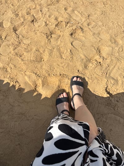 A person is sitting on sandy ground, viewed from above. The individual is wearing black sandals and a black and white patterned garment. The sand displays numerous footprints. In the lower left portion of the image, a shadow with a serrated edge is cast, suggesting the presence of an object like a fence or a lounger. The lighting indicates it is daytime. There is no indication of the specific location within Novaya Usman, Russia.