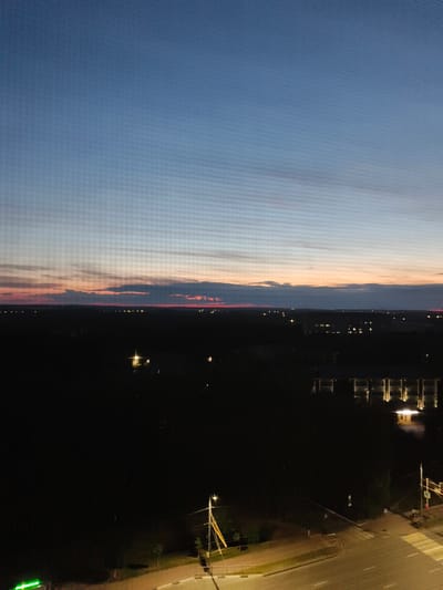 The image is a nighttime aerial view of a city at dusk, likely in Bryansk, Russia. The sky is a gradient of deep blue transitioning to lighter blues and then to shades of orange and pink near the horizon, indicating sunset or twilight. Wispy clouds are visible in the sky. Below the sky, a dark silhouette of a landscape stretches across the frame, punctuated by scattered lights from buildings and streetlights. In the foreground, a road with pedestrian crossings and streetlights is visible, along with a dark mass of trees and a building illuminated from the outside. The image appears to have been taken from a high vantage point, possibly a tall building or drone. The overall impression is one of a tranquil urban landscape transitioning into night.