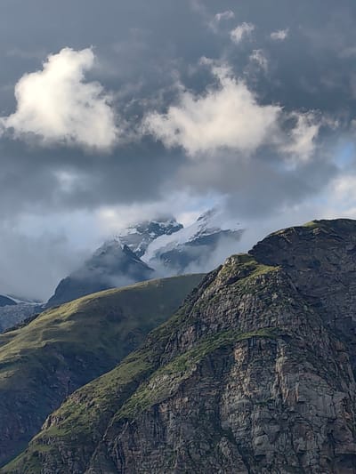 This image captures a mountainous landscape near Gramphu, India, under a dramatic sky. In the foreground, rugged, dark grey rock faces with patches of green vegetation rise steeply. Behind these, rolling green hills ascend towards higher peaks. The distant mountains are snow-capped and partially obscured by swirling grey clouds. The sky is a mix of dark, heavy storm clouds and patches of brighter, illuminated clouds, suggesting a dynamic and potentially turbulent weather system. There are no people or visible structures in the scene. The environment is characterized by raw, natural terrain.