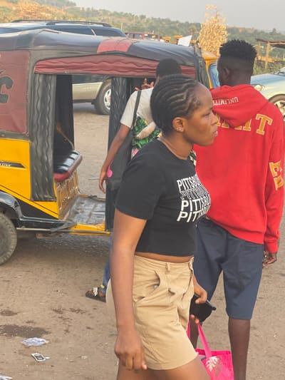 A woman with braided hair, wearing a black crop top and tan shorts, holds a cell phone. She stands near a yellow auto rickshaw with a brown and black design. Behind her, a man in a red sweatshirt and blue shorts is visible, along with another person partly obscured by the rickshaw. The setting appears to be a street scene with other vehicles and people in the background.