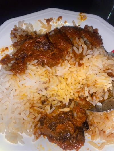 A close-up, top-down view captures a white plate with scalloped edges containing a meal, observed in Tudun Nupawa, Nigeria. The dish consists primarily of white, parboiled long-grain rice, largely covered by a reddish-brown, thick stew. Several dark, irregular pieces of meat or protein are visible within the stew, particularly concentrated towards the center and lower right. A silver-colored spoon is partially submerged into the food on the lower right side of the plate, indicating partial consumption or readiness for eating. The background is uniformly dark and out of focus, suggesting an indoor setting under low or artificial lighting conditions. No individuals are visible.
