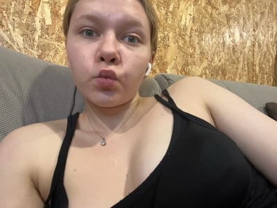 This is a portrait-style, slightly blurry, upside-down selfie of a young Caucasian woman with blonde hair and fair skin. She is wearing a black top with thin straps and a small silver necklace with a pendant. She also has white wireless earbuds in her ears. Her eyes are open and looking towards the camera, and her lips are pursed. The background consists of a textured grey surface and a wall made of oriented strand board (OSB), typical of unfinished interior spaces. The lighting appears to be artificial, suggesting an indoor setting. There is no discernible time of day or weather information. The image was taken in Klepiki, Russia. The woman's expression is neutral to playful.