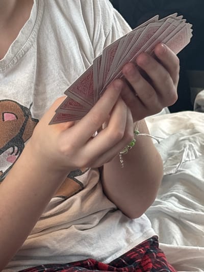 A close-up image, set in Barabinsk, Russia, depicts an individual's hands holding a fanned arrangement of approximately 15-20 playing cards. The cards feature a red background with a white, intricate design on their backs. The individual's left hand supports the fanned cards, while the right hand appears to be adjusting or holding the spread. The visible attire consists of a light grey or white t-shirt featuring a portion of a brown and pink graphic on the lower left. Below the t-shirt, a pattern of red and black plaid fabric is visible, consistent with trousers or blankets. A bracelet, comprising green beads and small silver-colored charms, is worn on the individual's right wrist. The immediate background shows wrinkled white fabric, suggestive of bed linen.