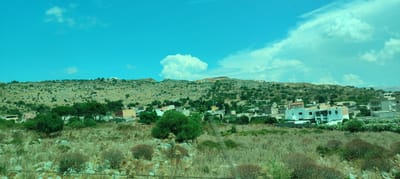 The image shows a rural landscape with a town nestled in a valley at the base of a rocky, scrub-covered hill. The sky is a vibrant turquoise with scattered white clouds, including a large, puffy cloud formation over the hill. The foreground consists of dry, yellowish-green grass and scattered rocks, with some low-lying bushes. In the middle ground, a cluster of buildings forms a small town. The buildings are a mix of colors, including white, light orange, and beige, with terracotta roofs visible on some. Some buildings have balconies and windows. Green trees, mostly tall and slender pines, are interspersed among the buildings and dot the landscape. The hill in the background is arid, with patches of green scrub and exposed rock. The overall impression is of a sunny, dry day in a Mediterranean setting. The image appears to have been taken from a moving vehicle, possibly a car or train, as evidenced by a faint horizontal line across the bottom of the frame.