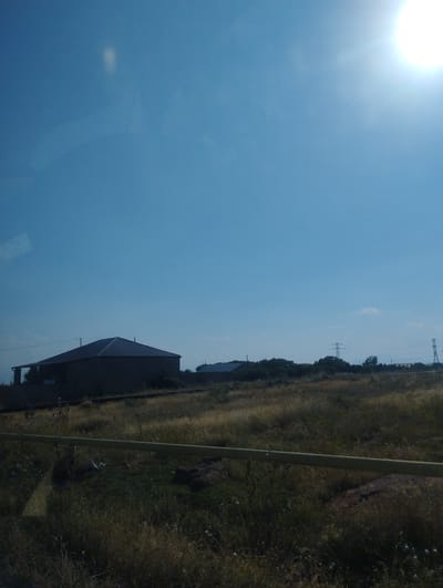 A wide shot captures a rural landscape under a bright, clear sky, with the sun high and slightly to the right, its rays creating a lens flare. The scene appears to be in Dashtadem, Armenia, given the context. In the foreground, a yellowish pipe runs horizontally across the frame, obscuring some of the dry, grassy terrain below. The middle ground features a dark, silhouetted building with a pitched roof, likely a house, situated on a slight incline. Further in the distance, a line of trees and vegetation can be seen, punctuated by power transmission towers. The overall impression is one of a sunny, open, and somewhat sparsely populated area.