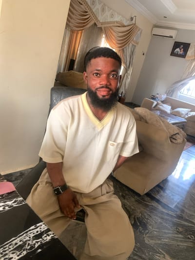 The scene depicts an interior residential setting, specifically a living room, located in Eika-Adagu New Layout, Nigeria. A male subject is seated, positioned slightly off-center, looking directly at the camera with a neutral to subtly smiling expression. He possesses a dark complexion, short dark hair, and a full dark beard. The subject wears a light cream-colored short-sleeved V-neck top with a light yellow trim and a small dark emblem on the left chest pocket. He is also wearing loose-fitting light brown trousers. A black wristwatch with a square buckle is visible on his left wrist.
The subject is seated in a dark-colored chair, with a dark, white-veined marble-patterned table partially visible in the left foreground. The background reveals a room furnished with light beige upholstered sofas and a dark, reflective coffee table. An ornate window treatment featuring multiple layers of light-colored curtains with decorative valances is present. A white wall-mounted split-unit air conditioner is affixed to the upper right wall, below which hangs a framed picture. The floor is composed of dark, polished tiles with lighter patterns, reflecting ambient light. The walls are light cream in color. The room is illuminated by natural light, likely from the visible window.