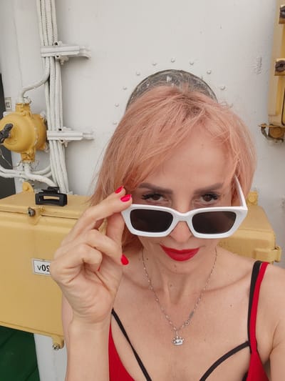 A close-up shot features a woman with peach-colored hair, wearing a red top and sunglasses. She is adjusting her white-framed sunglasses, revealing her eyes and red lipstick. A delicate silver necklace with a crown pendant rests on her collarbone. The background consists of a white wall with various industrial elements. To the left, there's a yellow box with a label that reads "VOS". White ropes are secured to the wall with white clamps. To the right, a yellow metallic object is attached to the wall. The lighting suggests daylight. The woman's expression is enigmatic, with her gaze directed at the viewer. The setting appears to be outdoors, possibly on a ship or a pier, given the presence of nautical-like equipment. The image conveys a sense of casual confidence and style.