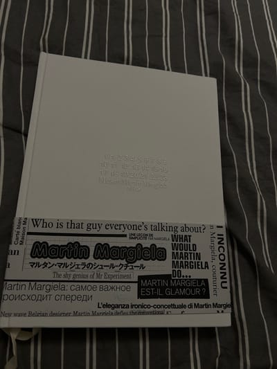 The image shows a book laid on a striped surface. The book's spine has vertical numbers from 0 to 23 and the text "Maison Martin Margiela Paris". The front cover is a collage of text snippets relating to Martin Margiela. These include phrases like "Who is that guy everyone's talking about?", "Martin Margiela", "What Would Martin Margiela Do...", and quotes in other languages. A small ribbon bookmark is visible.