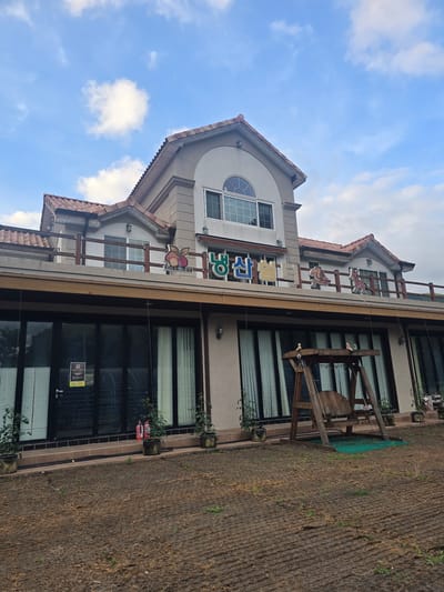 The image depicts a two-story building with a tiled roof, situated in Gumi-si, South Korea. The building has a modern architectural style with a light-colored facade, large glass windows on the ground floor, and a balcony on the second floor. A wooden swing set is positioned in front of the building. The ground in front of the building appears to be recently prepared for landscaping, with rows of brown material and sparse vegetation. The sky above is partly cloudy. A sign with Korean characters and cartoonish figures is visible on the second-floor balcony railing. The overall scene suggests a commercial establishment, possibly a cafe or restaurant, with an outdoor seating area.