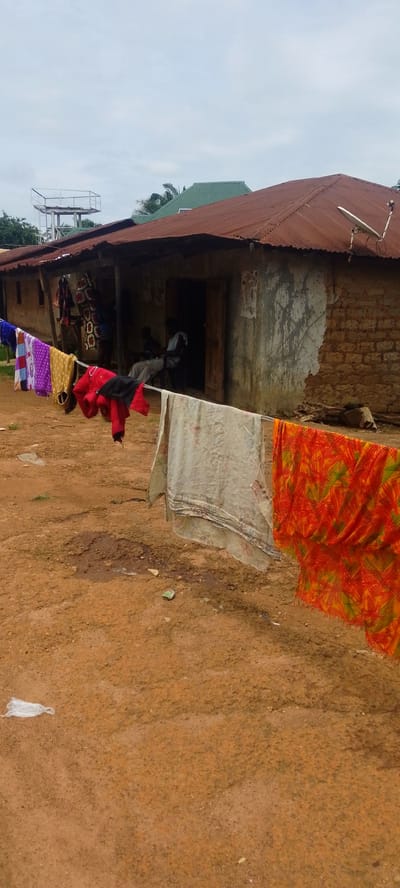 The image shows a modest, rustic dwelling in Baissa, Nigeria, with a corrugated metal roof that has seen better days. In front of the house, laundry hangs on a clothesline, including colorful patterned fabrics and a red jacket. The ground in front of the house is a reddish-brown dirt, with some patches of wetness and scattered debris. The building itself appears to be constructed from mud bricks with some sections plastered. A doorway leads into the dark interior of the house, where two people are seated. One is a man wearing a white shirt and light-colored pants, and the other is partially visible, dressed in darker clothing.

Above the house, a metal water tower with a railing is visible, suggesting the presence of a water supply system. The sky is overcast, indicating a cloudy or possibly rainy day. The overall scene conveys a sense of everyday life in a rural setting, with the focus on basic necessities like shelter and clean clothes. There is no visible text in the image.