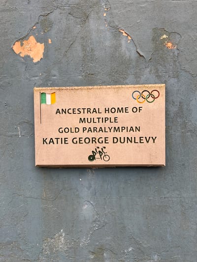 A stone plaque is affixed to a textured, dark grey wall with peeling orange paint in the upper left quadrant. The plaque features carved text and imagery. At the top left of the plaque, an Irish flag is depicted. To the right of the flag, the Olympic rings are carved and colored in their traditional blue, yellow, green, red, and black.
Below the flag and Olympic rings, carved text reads: "ANCESTRAL HOME OF MULTIPLE GOLD PARALYMPIAN KATIE GEORGE DUNLEVY". Beneath the text, a stylized carved image depicts a person on a bicycle. The plaque is a light brown, sandy color and appears to be made of stone or a similar material. The wall surrounding the plaque is rough and uneven. The overall scene is an outdoor public space, likely a building facade. The location is identified as Mountcharles, Ireland.