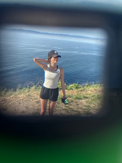 A young female is observed through an optical device's view, framed by dark, soft-edged borders. The scene is set on a grassy cliffside in Palos Verdes Estates, United States, overlooking the Pacific Ocean.

The female, appearing as a young adult, stands with her body angled slightly to the right, her head turned to gaze leftward across the ocean horizon. She wears a dark baseball cap with a light-colored logo, a light grey tank top, and black athletic shorts. Her right arm is raised with the hand positioned behind her neck, while her left hand holds a dark object with green accents, possibly a water bottle or small container.

The immediate foreground consists of dry, sparse grass and light green vegetation on a sloping cliff. A wide, calm blue ocean dominates the midground, its surface showing subtle ripples. Hazy mountain ranges or distant landmasses are visible on the far horizon under a clear, light blue sky. The scene is illuminated by bright daylight.