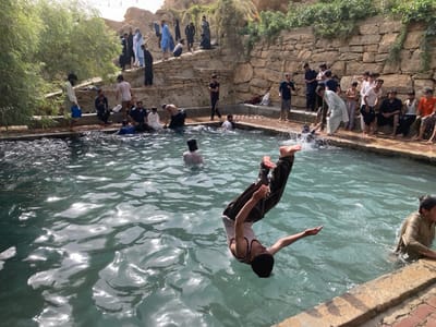 This image depicts an outdoor recreational scene at a natural water pool in Killi Malik Sher Mohammad, Pakistan. A male subject, appearing to be a young adult, is captured mid-air performing a backward flip into the turquoise-colored water. He wears dark trousers and a light-colored sleeveless top, with water visibly splashing from his entry.
The pool is an elongated, rectangular structure, partially lined with cut stone blocks, and its surface reflects sunlight. Approximately thirty to forty males, predominantly teenagers and young adults, are present in and around the pool. Several individuals are partially submerged in the water, while a larger group congregates on the stone banks and on elevated, terraced rocky areas behind the pool. Most individuals on the banks are observing the activity in the water.
Clothing varies, with many males wearing traditional Pakistani shalwar kameez in colors such as white, black, and light blue. Others wear casual shirts and trousers. One individual on the left bank holds a blue bucket. The background consists of arid, rocky hillsides with sparse green vegetation, including a dense cluster of leafy trees on the left side. The scene is illuminated by bright daylight.