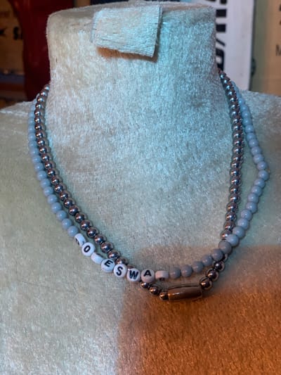 A close-up image of a silver and light blue beaded necklace. The necklace is made of two strands of beads, one silver and one light blue. The silver beads are round and metallic, while the light blue beads are slightly oval-shaped and have a matte finish. The beads are strung together with a silver metal thread. The necklace has a silver clasp and is lying on a light gray, fuzzy surface. The necklace spells out the word "SWAG" using white letter beads. The image was taken indoors, likely in a home or shop. The coordinates provided (latitude: 7.282, longitude: 5.161) are in the city of Ita Oniyan, Nigeria. There are no people or actions in the image.