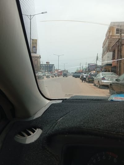 The image is taken from inside a car, looking out through the windshield onto a street in Benin City, Nigeria. The road is lined with parked cars on the right side. A few pedestrians are walking on the street. There are buildings on both sides of the street, including a business with a sign that reads "ELECTRONIC WAREHOUSE at WHOLESALE Prices." The sky is overcast. The car's dashboard is visible, including a section of the speedometer and an air vent. The windshield appears to have a crack.