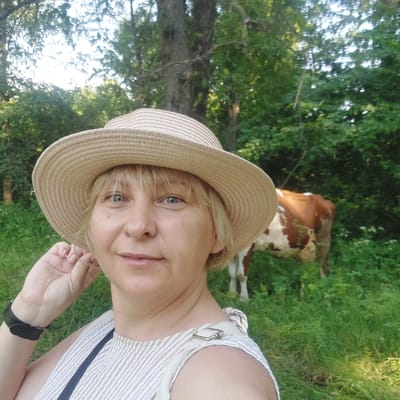 A selfie is taken by a fair-skinned woman wearing a straw hat and a light-colored, pinstriped dress. She has blonde hair and is looking to her right with a slight smile. Her right hand is raised to adjust her hat. She is wearing a black watch on her left wrist. In the background, a brown and white cow is visible standing in a grassy area with trees. The scene appears to be outdoors in a rural or park-like setting, possibly on the outskirts of Voronkiv, Ukraine, judging by the surrounding vegetation and the presence of livestock. The lighting suggests it is daytime.