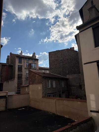 This image presents an urban back alley or courtyard scene in Saint-Étienne, France, captured under a partly cloudy sky with bright sunlight.
In the background, a multi-story residential building with a light-colored, possibly stucco, facade and a pitched roof is visible, featuring multiple windows with dark frames and several chimneys. To its immediate right, a taller, windowless building section composed of dark, irregular stone masonry rises prominently.
In the mid-ground, a two-story house with a reddish-brown tiled roof, light-colored walls, and multiple windows (some with white shutters) is situated. To its right, a darker building wall with some vertical plant growth is partially obscured.
The foreground consists of a dark, flat, paved area, likely asphalt, surrounded by a low, light beige-colored wall topped with terracotta coping tiles. The extreme right edge of the frame shows a cream-colored building facade with dark trim and a dark, closed roller shutter on a window. No individuals or active subjects are present in the scene.