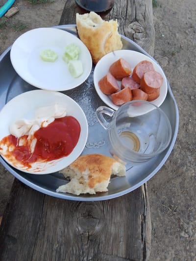 A silver tray holds a rustic outdoor meal. The tray contains a white plate with sliced cucumbers, a white bowl with sliced sausages, and another white bowl with a dollop of mayonnaise and a generous portion of ketchup. Pieces of bread, torn by hand, are scattered across the tray and to the side, with one larger piece resting on the tray. An empty glass mug with a handle sits on the tray, its interior showing some faint residue. The tray itself rests on a weathered wooden surface, suggesting an outdoor setting. The ground around the wooden surface is a mix of dirt and sparse vegetation, with some dry twigs and small stones visible. The lighting suggests it is daytime, likely outdoors in a casual or rustic environment. There is no visible text in the image. The overall impression is of a simple, informal meal being enjoyed outdoors. The location context of Кугейский, Russia, further reinforces this rustic outdoor setting.