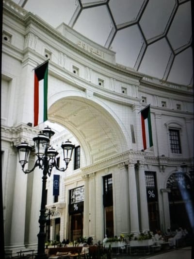 The image shows the exterior of a grand, white building with a distinctive honeycomb-patterned glass roof. The building features classical architectural elements, including columns and decorative moldings. Two national flags, the flag of Kuwait, are prominently displayed, one on each side of a large arched entryway. In the foreground, a decorative black lamppost with multiple lamps stands to the left. The building's facade includes several windows and entrances, some with businesses visible behind them, indicated by signage with percentage symbols. A group of people are seated at tables outside one of the establishments, suggesting a public space or commercial area. The overall scene evokes an impression of a modern, well-maintained urban setting, likely a public building or a complex of shops and restaurants. The location is identified as Golbembal, Nigeria, though the architectural style does not strongly suggest a Nigerian context, and further information would be needed to confirm this locale.