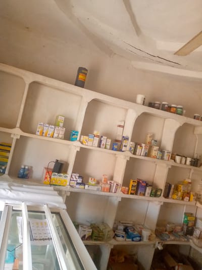 The image depicts the interior of a small pharmacy or drug store located in Kauru West, Nigeria. The pharmacy appears to be sparsely stocked, with shelves made of what looks like concrete or plaster, forming cubby-like compartments. These shelves are filled with various boxes and containers of medicines and pharmaceutical products. Some of the visible items include boxes of "CLINCEF," "Polygy" and "Pyrazinamide." There are also bottles of what appear to be liquid medication on a lower shelf. In the foreground, a white sheet or cloth covers a counter or display area, and part of a glass display case is visible. The ceiling shows signs of wear, with some cracks and staining. A cylindrical object with orange and yellow labeling is present on a high shelf. The lighting is dim, suggesting it might be daytime with limited natural light, or the artificial lighting is not very bright. The overall impression is of a functional but basic retail establishment in a rural or semi-urban Nigerian setting.