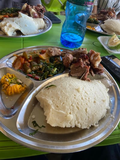A stainless steel plate with four sections contains a meal. In the top section, a large portion of white, soft-looking ugali (cornmeal porridge), is placed next to a section with a stew of green leafy vegetables, okra, tomatoes, and onions, and a separate section with a smaller portion of ugali and a side of yellow corn kernels. The last section contains a large portion of meat, likely goat or beef, that is brown and looks like it was braised. The plate is on a bright green surface, and a blue plastic water bottle is on the right side of the plate. The location of the photograph is in Dodoma, Tanzania at coordinates -6.161, 35.764.